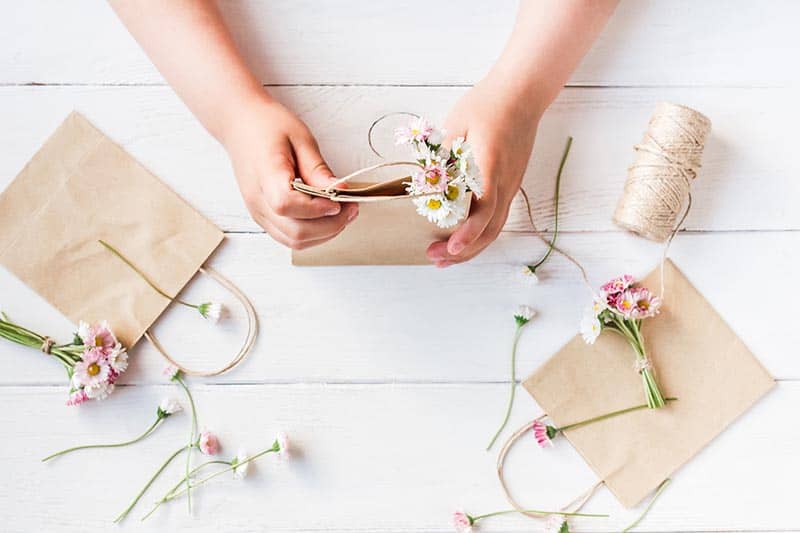 image of a kid's hands folding dried flowers into a gift bag. Art supplies surround the hands.
