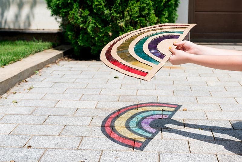 image of a rainbow prism art project, held by kids' hands and reflecting onto a driveway